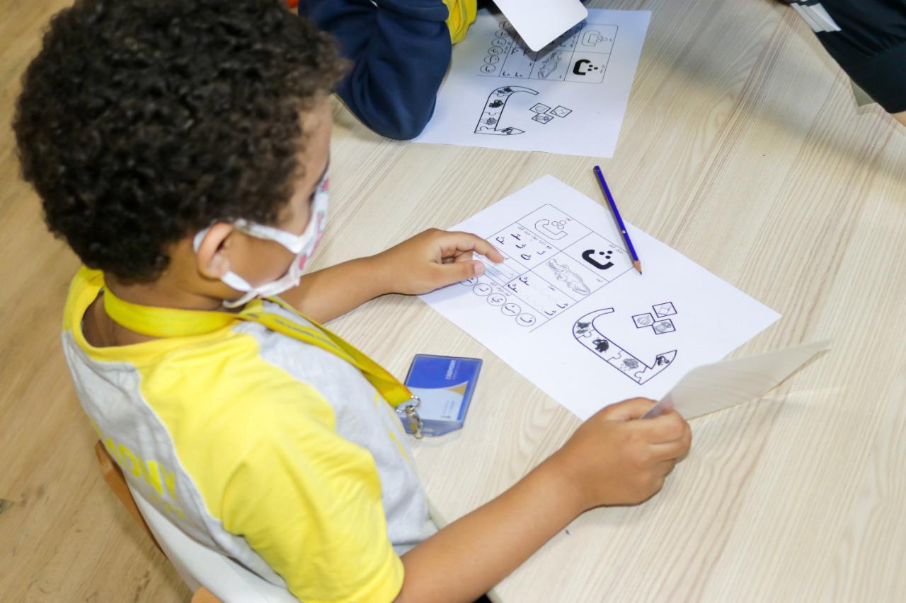 A child at IVY STEM International School actively engages in educational activities while following health precautions during the COVID-19 pandemic. The child wears a face mask, holds a pencil, and points to a spot on a paper filled with learning exercises.